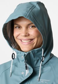 Vattentät turkosa regnjacka med dragsko i huvan, dragkedja fram och silverdetaljer. Logotyp synlig på vänster framsida. Slät yta.