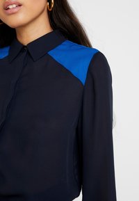 Blusa de un oscuro color marino con acento azul en el hombro, que presenta un cuello clásico y mangas largas. Tejido suave con un ligero brillo.