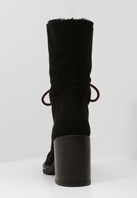 Black suede ankle boots with a high, chunky heel, featuring a fuzzy top and decorative red laces on the sides.