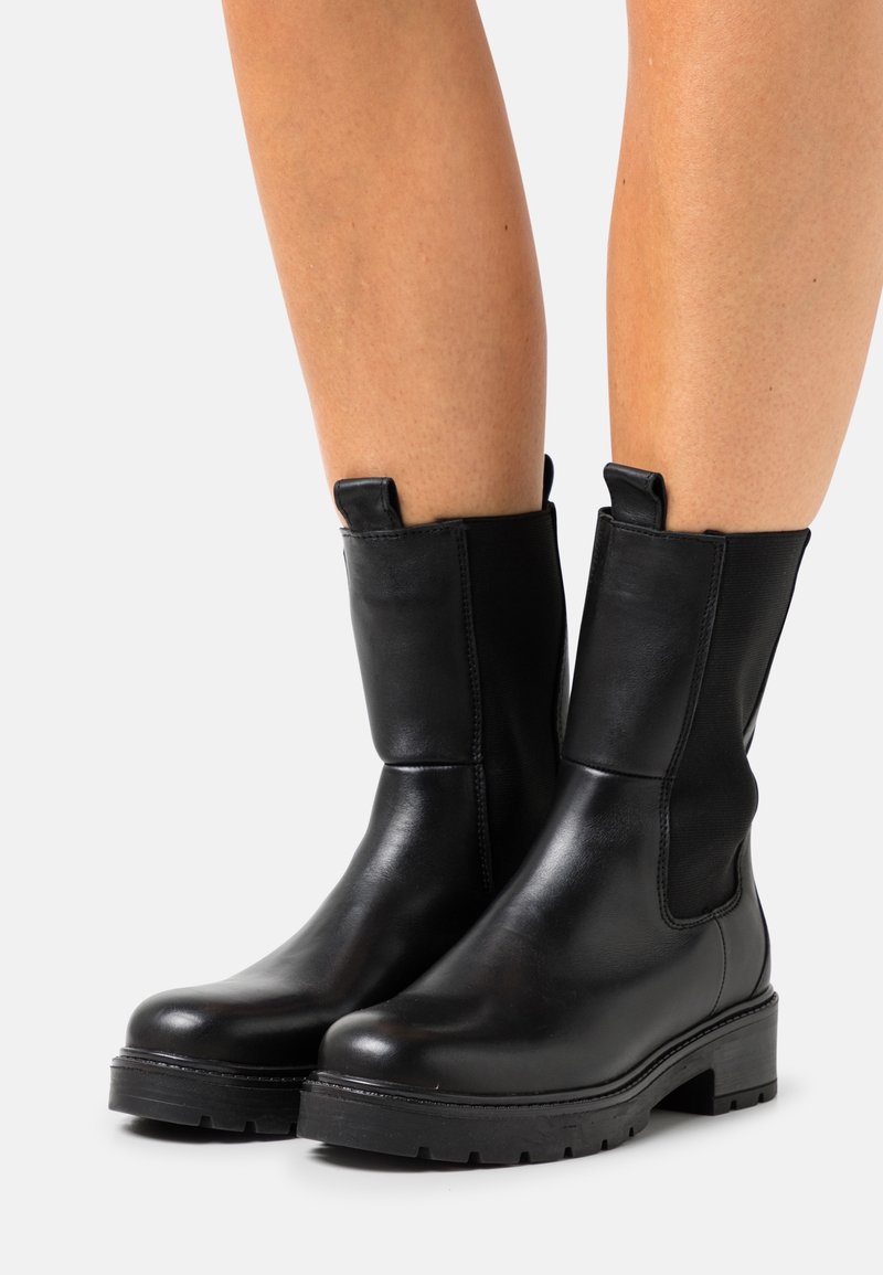 Bottes en cuir noires mi-mollet avec semelles épaisses portées sur jambes nues, dotées de panneaux élastiques sur les côtés et de languettes de tirage en haut.