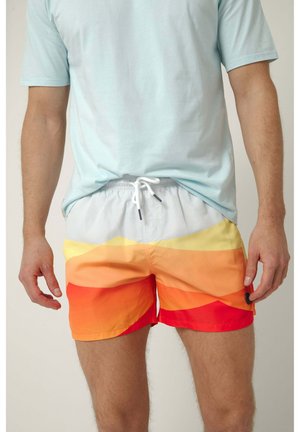 Hombre con camiseta azul claro y pantalones cortos de baño coloridos con un patrón degradado en blanco, amarillo, naranja y rojo, de pie frente a un fondo neutro.