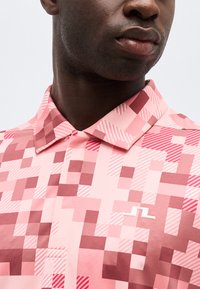 Κοντινό πλάνο ενός ατόμου που φοράει ροζ πουκάμισο με ψηφιακό μοτίβο και γιακά, με ένα μικρό λευκό λογότυπο στο στήθος.