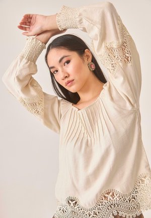 Blusa de color crema con mangas largas y voluminosas. Presenta detalles fruncidos, delicados acentos de encaje en las mangas y el dobladillo, y un escote cuadrado.