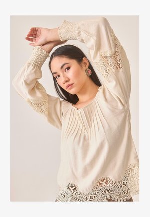 Blusa de color crema con mangas largas y voluminosas. Presenta detalles fruncidos, delicados acentos de encaje en las mangas y el dobladillo, y un escote cuadrado.