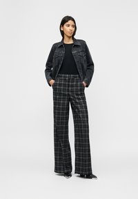 Γυναίκα στέκεται με τα χέρια στις τσέπες φορώντας μαύρο τζιν μπουφάν, μαύρο τοπ, φαρδιά γκρι παντελόνια με καρο μοτίβο και μαύρες μπότες με ασημένιες μυτερές λεπτομέρειες.