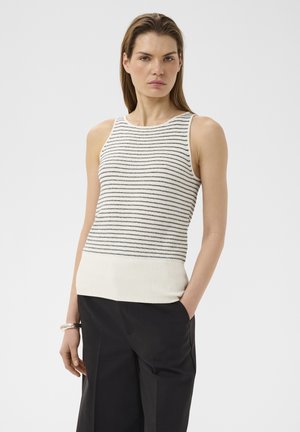 Kobieta w białym bezrękawniku z cienkimi poziomymi czarnymi paskami i czarnych spodniach, stojąca z jedną ręką w kieszeni na białym tle.