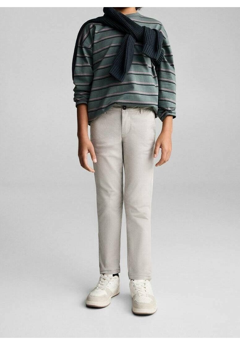 Enfant portant un t-shirt à manches longues rayé gris avec un pull foncé posé sur les épaules, un pantalon beige et des baskets blanches, debout sur un fond gris.