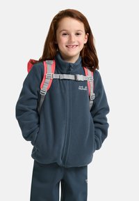 Fleecejacke in Dunkelblau mit Reißverschluss, ausgestattet mit einem hohen Kragen und Logo. Das Modell trägt einen Rucksack mit pinken Trägern und grauem Zubehör.