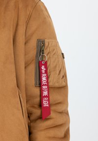 Chaqueta de ante marrón con un bolsillo con cremallera en la manga izquierda, que presenta una etiqueta roja "RETIRAR ANTES DEL VUELO" adjunta.