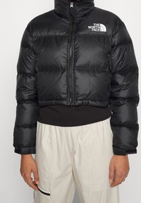 Korta svarta pufferjacka i glänsande material, med hög krage, framdragkedja och vitt logotyp på vänster bröst.