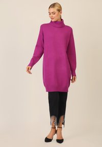 Übergroßer lila Rollkragenpullover mit gerippten Bündchen und Saum. Kombiniert mit schwarzen Jeanshosen mit fransigen, verzierten Kanten. Schwarze Absatzschuhe.
