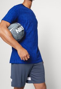 Královsky modré žebrované sportovní tričko spárované se šedými šortkami, držící šedý fotbalový míč Nike s výrazným logem.