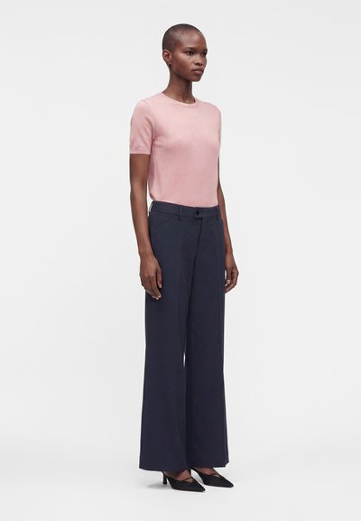 Femme debout portant un haut rose clair à manches courtes, un pantalon large bleu marine et des talons noirs pointus sur un fond blanc uni.