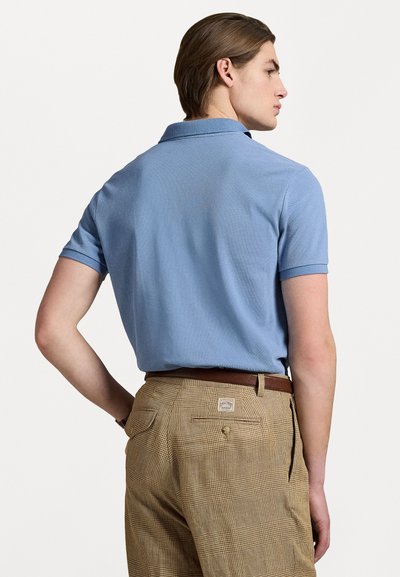 Camisa polo azul claro con tejido texturizado, mangas cortas y cuello; combinada con pantalones cuadros marrones que presentan un detalle de bolsillo trasero.