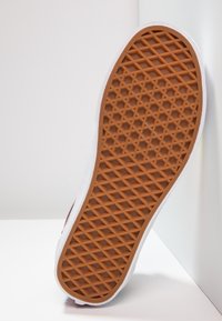 Semelle de chaussure avec motif gaufré marron et bord blanc, inclinée contre une surface blanche d'angle.