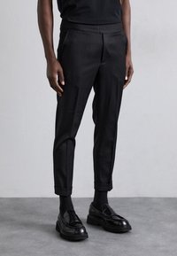 Pantalon noir sur mesure avec une taille élastique, coupe slim et revers roulés, associé à des mocassins noirs et des chaussettes foncées. Texture lisse.