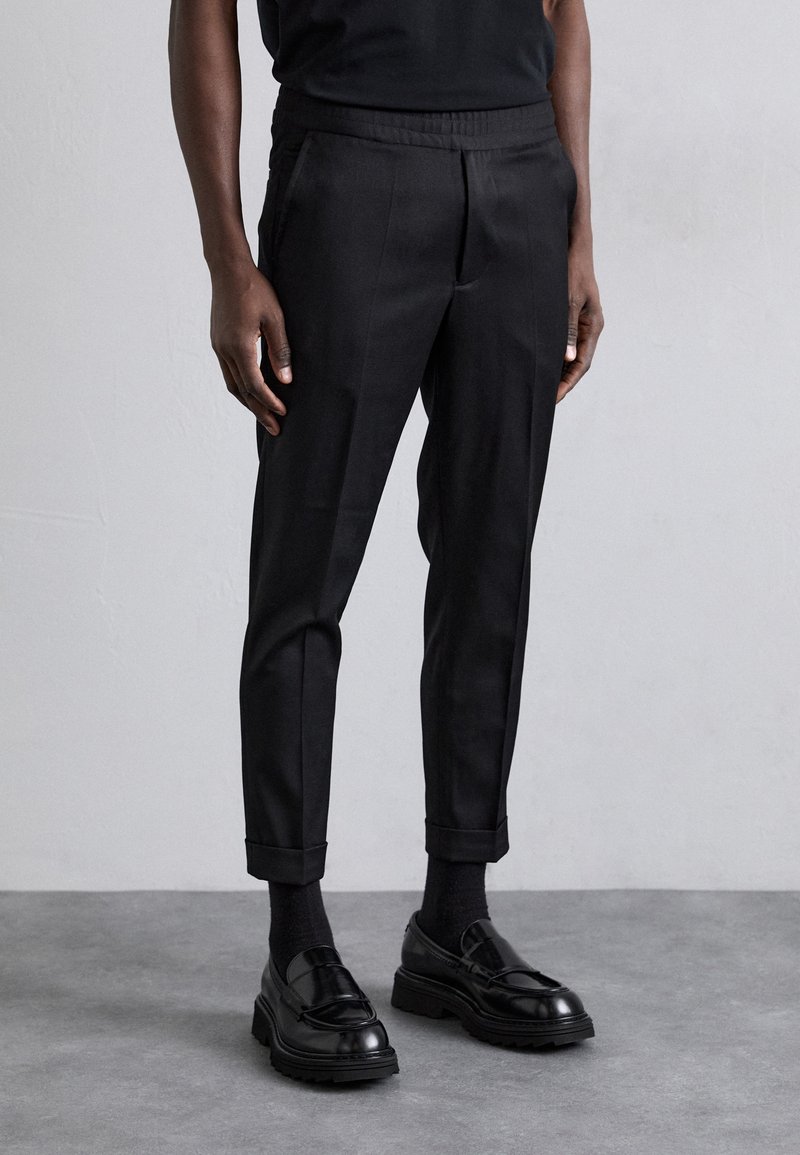 Pantalon noir sur mesure avec une taille élastique, coupe slim et revers roulés, associé à des mocassins noirs et des chaussettes foncées. Texture lisse.