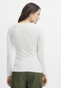 Langärmliges, weißes Rippoberteil mit weitem Ausschnitt, figurbetontem Design und glatter Textur, kombiniert mit olivgrünen Hosen.
