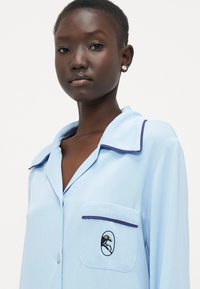 Cămașă de pijama de culoare albastru deschis cu un buzunar prezentând un design brodat. Are tiv bleumarin la guler și buzunar, realizată dintr-un material neted.