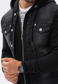 Homme portant une veste en jean noire par-dessus un sweat à capuche noir avec un t-shirt blanc à motif graphique, les mains jointes et portant une montre noire au poignet gauche.