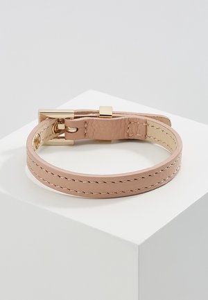 Lysebrunt læderarmbånd med guldbeslag og syningsdetalje, hvilende på en hvid piedestal mod en grå baggrund.