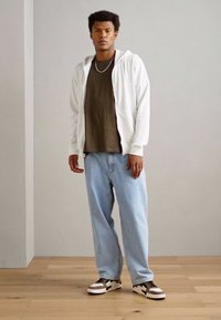 Bijela zip-up hoodie preko maslinasto zelene majice, u kombinaciji s svijetloplavim širokim trapericama i crno-bijelim tenisicama. Pod od drva.