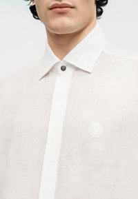 Κοντινή λήψη ενός ατόμου που φοράει λευκή λινή πουκάμισο με μυτερό γιακά και διακοσμητικό κουμπί στην κορυφή.