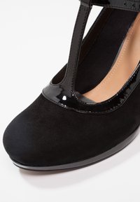 Chaussure noire en daim à bout rond avec bride en T vernie brillante et doublure intérieure beige sur fond blanc.