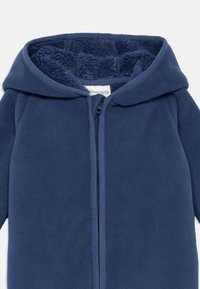 Combinaison en polaire bleu foncé avec capuche, fermeture éclair sur le devant et doublure intérieure douce et texturée. Finition extérieure lisse avec coutures ajustées.