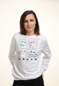 Sudadera blanca de manga larga que presenta un gráfico con cuatro paneles de un personaje con orejas grandes, acompañado del texto "GROGU" en azul.