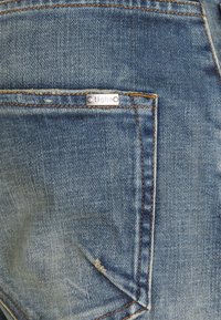 Dżinsy w kolorze wyblakłego niebieskiego, z pomarańczowymi przeszyciami i małą srebrną metką na tylnej kieszeni. Tekstura jest gładka z widocznym splotem.