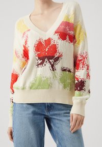 Pull en tricot avec un col en V, ornée d'un motif floral rouge, jaune et vert sur fond beige. Poignets et ourlet côtelés.