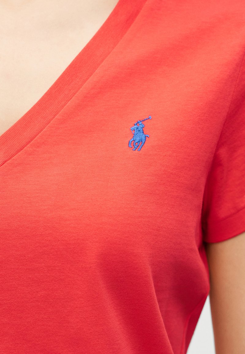 Close-up van een rood V-hals shirt met een klein blauw geborduurd polospelerlogo op de borst, gedragen door een persoon.