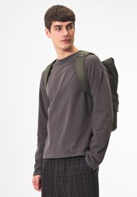 KHAKI Rucksack mit glatter Textur und verstellbaren Trägern, getragen über einem langärmligen grauen Hemd und karierter Hose, auf einem neutralen Hintergrund gezeigt.