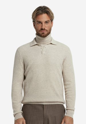 Beżowa swetrowa koszulka polo z wysokim kołnierzem, zapięciem na trzy guziki i długimi rękawami, połączona z brązowymi spodniami.