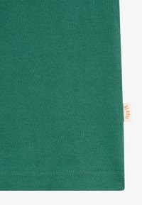Gros plan sur un tissu vert montrant une petite étiquette beige avec du texte orange "BLKVUS" cousue dans la couture sur le côté droit.