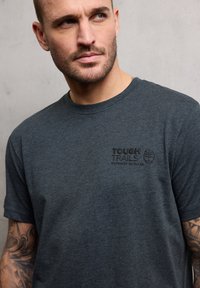 Tmavě šedé bavlněné tričko s kulatým výstřihem a krátkými rukávy. Na levé straně přední části je texturované logo s nápisem "TOUGH TRAILS" a motivem stromu.