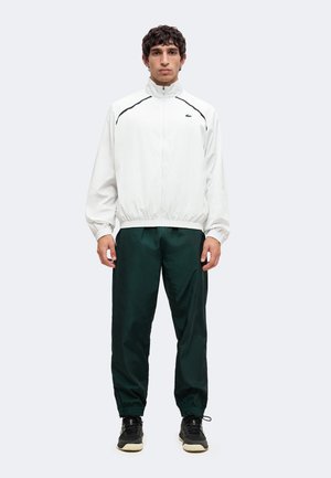 Mann trägt eine weiße Jacke mit Reißverschluss, dunkelgrüne Hose und schwarze Sneaker, steht frontal vor einem schlichten Hintergrund.