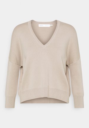 Beżowy sweter z dekoltem w serek, długim rękawem, ściągaczami na rękawach i dole, o luźnym, lekko skróconym kroju.