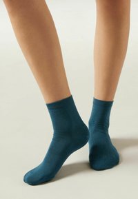 Calcetines de algodón en color azul verdoso con un puño acanalado y textura suave, de longitud hasta el tobillo, colocados sobre un fondo neutral.