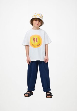 Παιδί στέκεται φορώντας ένα άσπρο μπλουζάκι με κίτρινο χαρούμενο πρόσωπο, μπλε παντελόνι, μαύρα σανδάλια και ένα άσπρο καπέλο με χαρούμενα πρόσωπα.