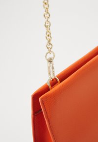 Borsa a tracolla arancione con texture, dotata di una catena dorata e gancio, che presenta un design liscio e strutturato con una forma angolare.