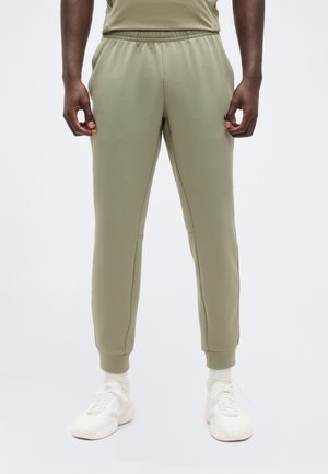 Homme portant un pantalon de jogging vert olive clair, des chaussettes blanches et des chaussures de sport blanches, debout devant un fond clair uni.