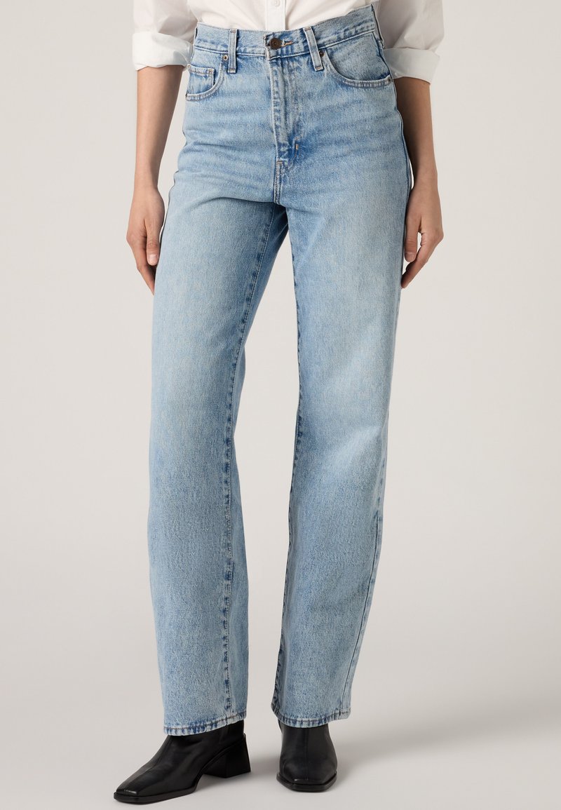 Person iført lyseblå højtaljede jeans med lige ben kombineret med sorte ankelstøvler og en hvid skjorte med ærmer rullet op.