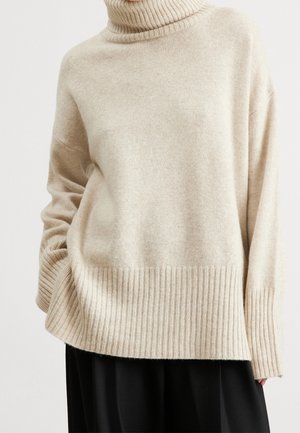 Beiger, übergroßer Rollkragenpullover mit gerippten Bündchen und Saum, getragen über einer schwarzen Hose, zeigt eine lockere Passform und weiche Textur.
