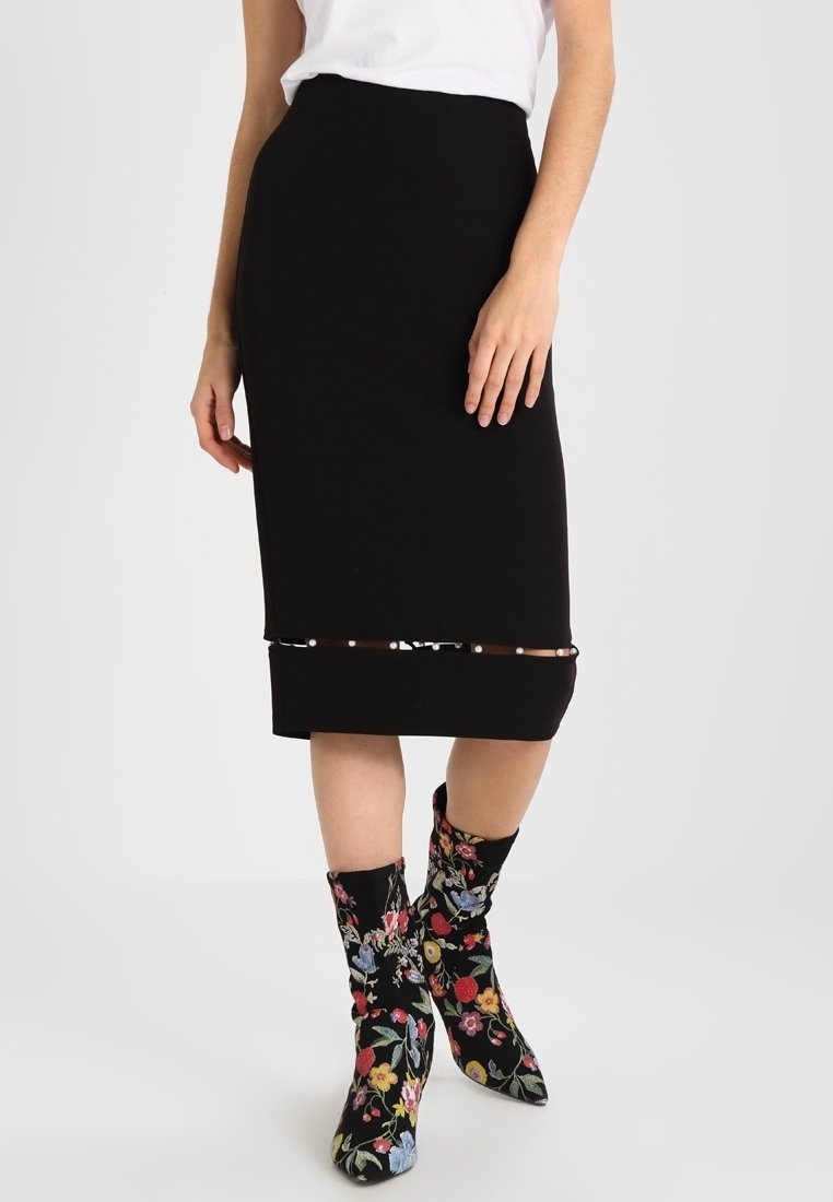 Femme portant une jupe noire longueur genou avec un ourlet transparent et des bottines à motifs floraux, debout devant un fond blanc uni.