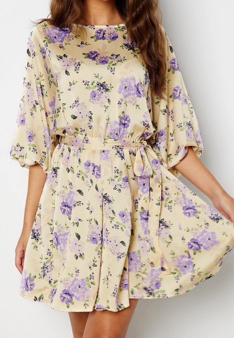 Femme portant une robe beige à imprimé floral violet, manches trois-quarts bouffantes et ceinture nouée à la taille, tenant la jupe écartée.