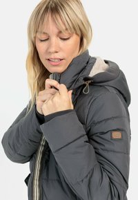 Grijze gevoerde jas met capuchon, fleecevoering en een ritssluiting aan de voorkant. Heeft ribbels aan de mouwen en een geweven label op de linkermouw.