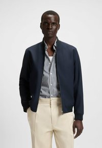 Navy bomber jacket with a zip front, ribbed cuffs, and a smooth texture, layered over a white and navy striped shirt, paired with cream trousers.
