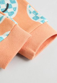 Oranges Baumwollstoff mit strukturiertem Finish. Besitzt blaue gemusterte Akzente an den Manschetten, umgeschlagenen Säumen und weichen, elastischen Kanten.